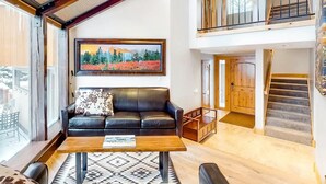 Ein geräumiges Wohnzimmer mit einem schwarzen Ledersofa, einem Holztisch und einem großen Landschaftsbild. Eine Treppe führt zu einer oberen Ebene mit einem Balkon.