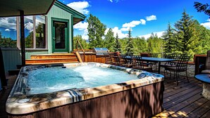 Ein Whirlpool auf einer Holzterrasse mit Gartenmöbeln und Blick auf Bäume und Berge.