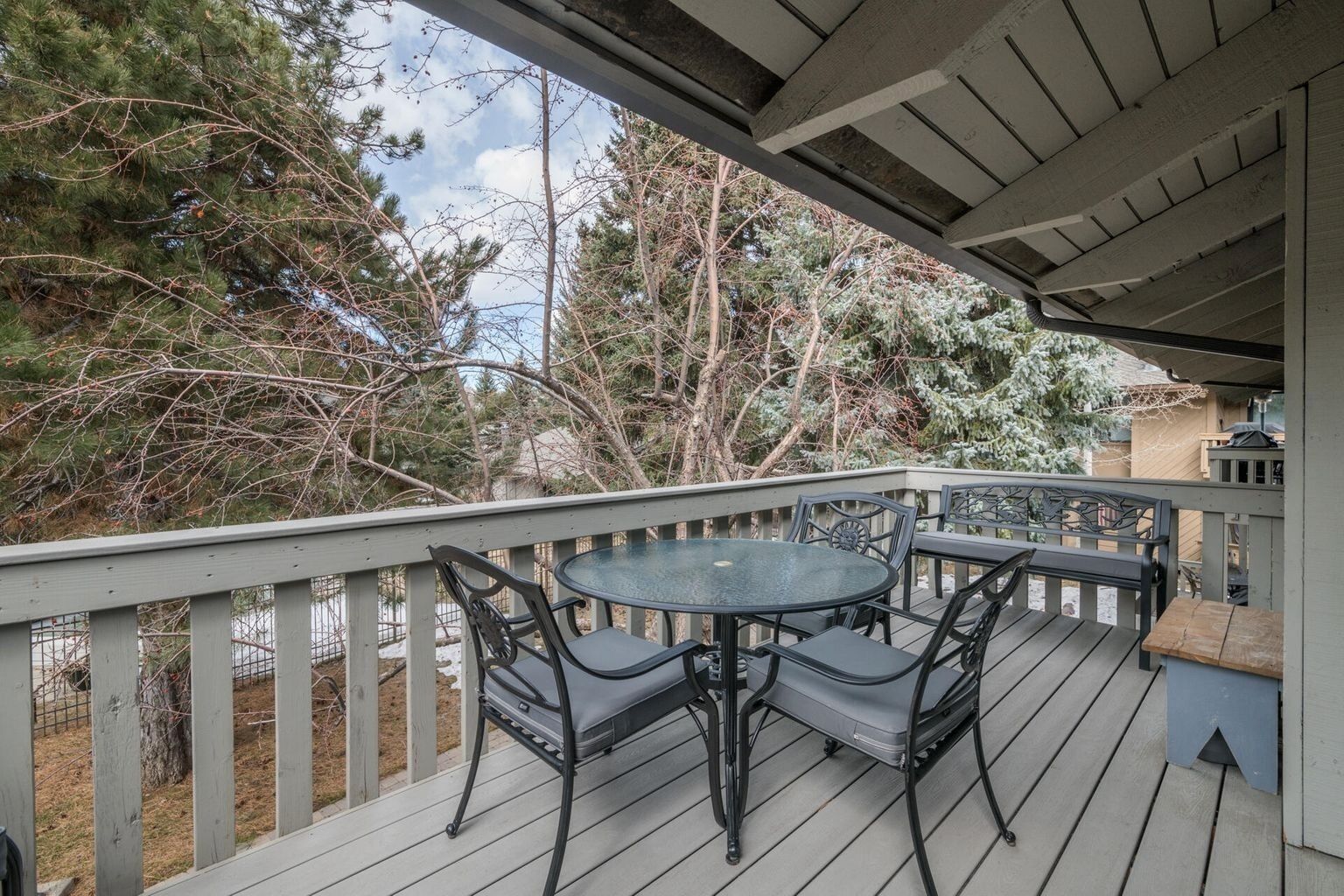 A covered outdoor deck with a round table and chairs, surrounded by a wooden railing and trees.