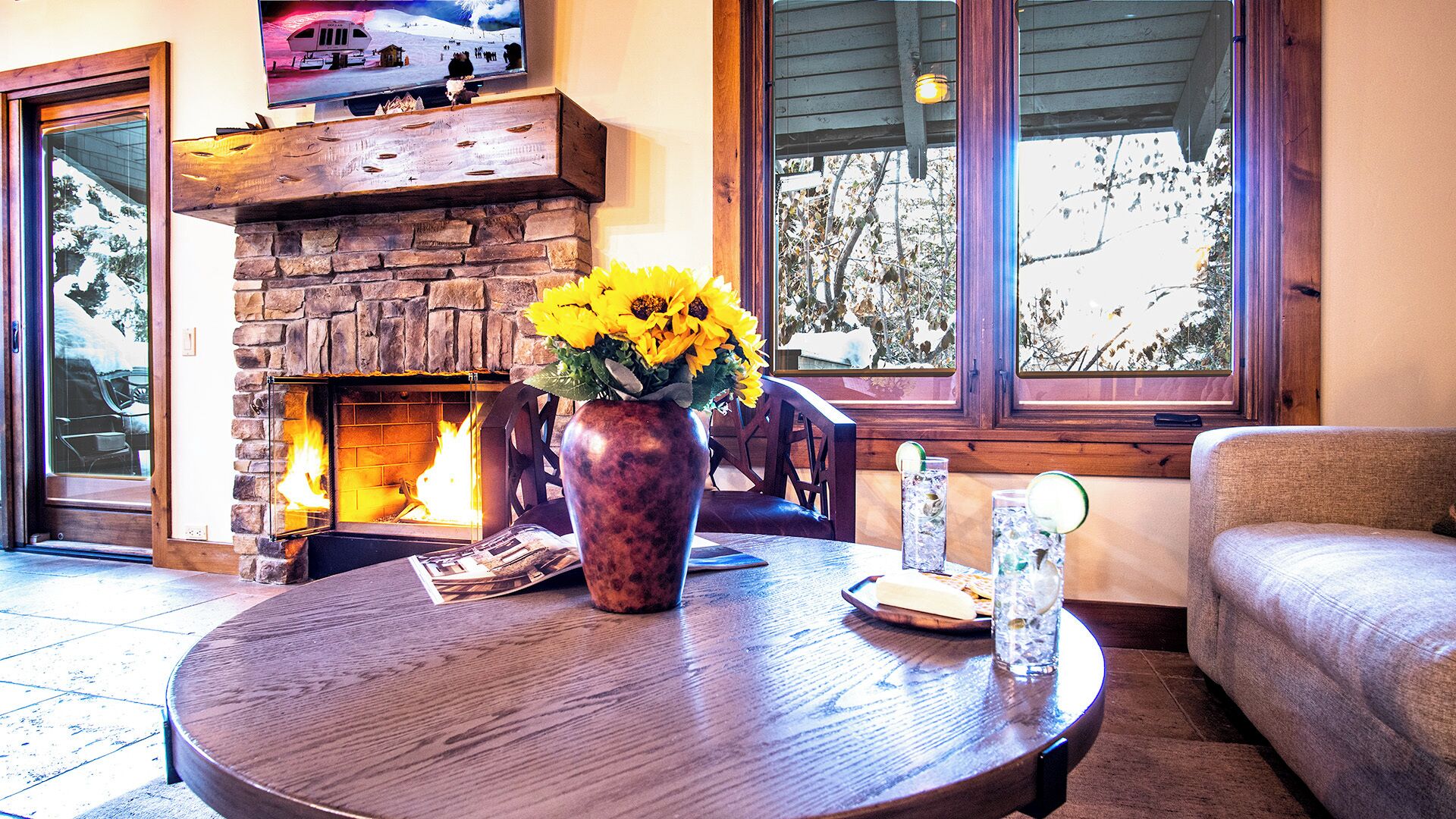 A cozy living room with a stone fireplace, a round wooden table with a vase of sunflowers, and a view of trees through large windows.