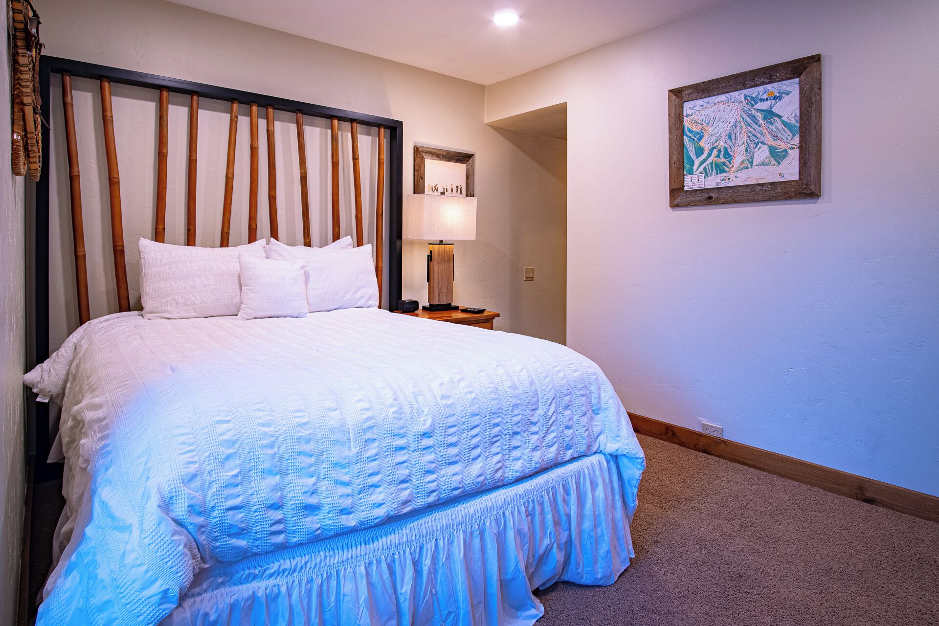 Una cama bien hecha con ropa de cama blanca y un cabecero de madera, una mesita de noche con una lámpara, y un mapa enmarcado en la pared.