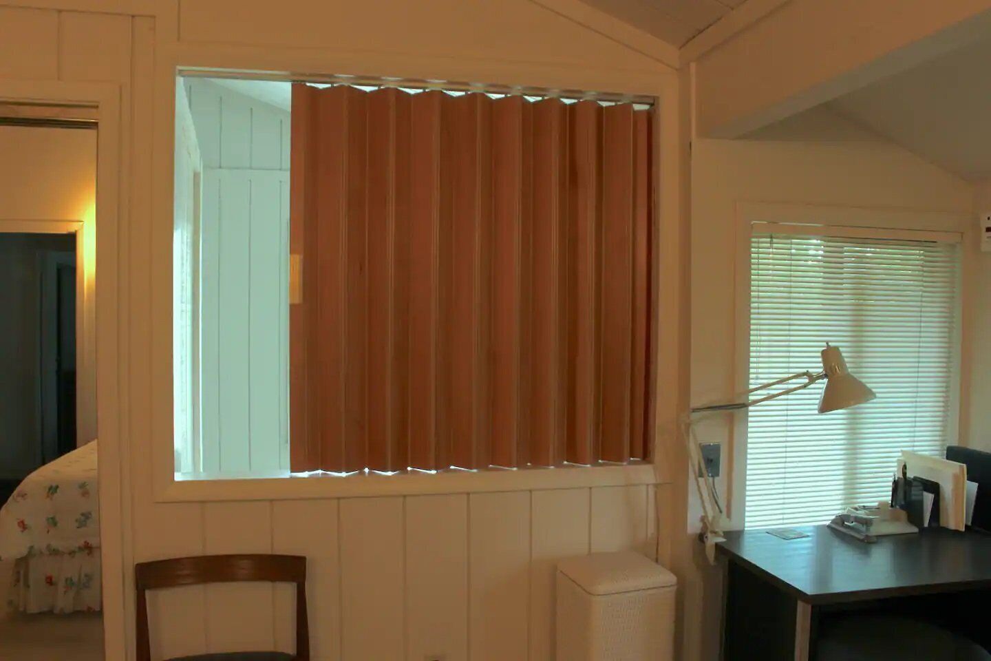 A room with a window covered by brown curtains, a desk with a lamp, and a chair.