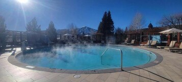 Una piscina circular con un centro azul cristalino, rodeada de sillas de descanso y sombrillas, y una montaña al fondo.