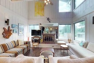 A spacious living room with a fireplace, large windows, and a high ceiling.