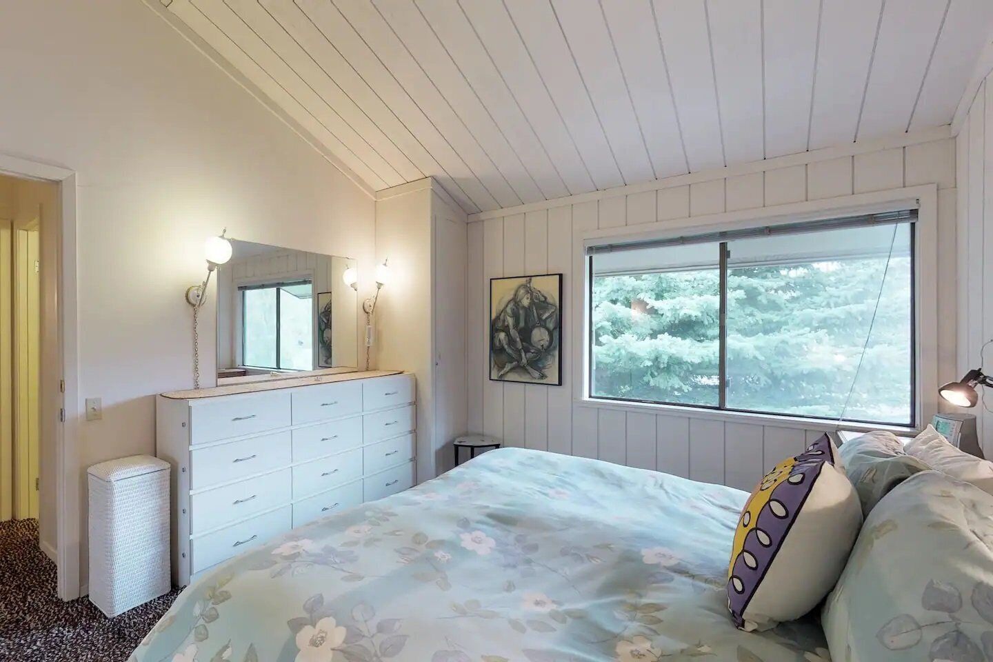 A bedroom with a large bed, a dresser, a window with a view of trees, and a mirror with lights.