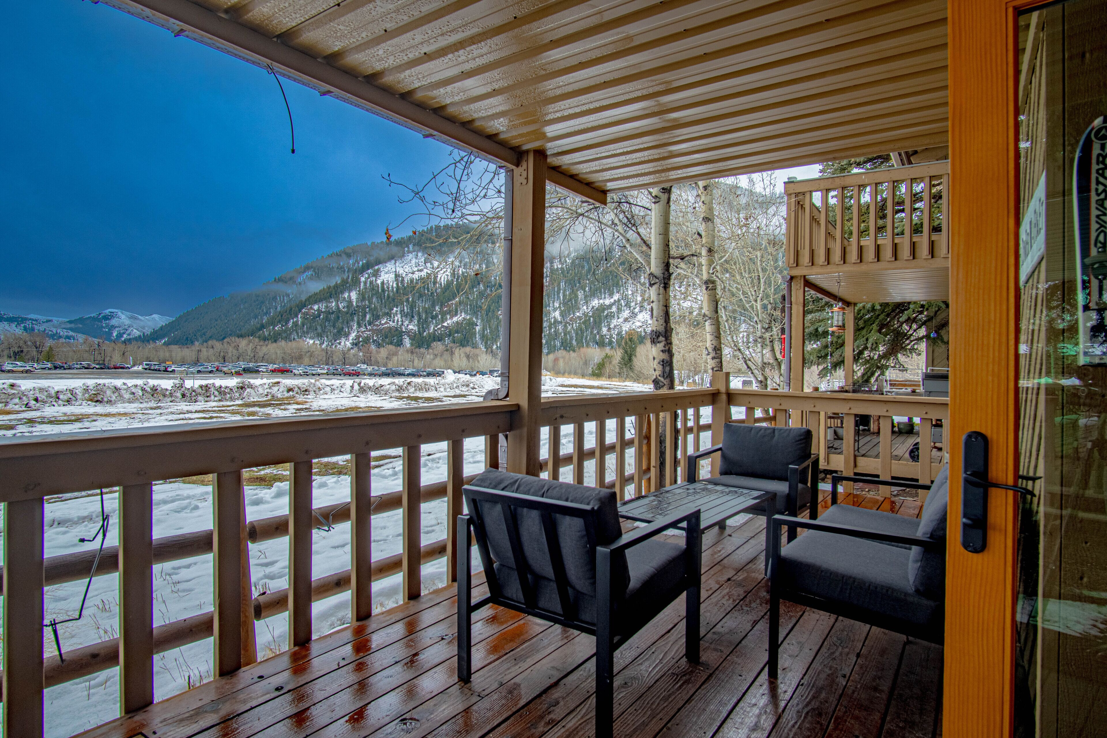 A wooden deck with two chairs and a table, overlooking a snowy landscape with mountains.