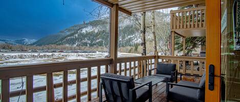 A wooden deck with two chairs and a table, overlooking a snowy landscape with mountains.