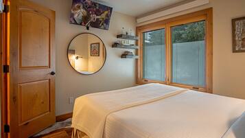 Ein Schlafzimmer mit einem großen Bett, einer Holztür, einem runden Spiegel, Regalen mit Flaschen und einem Fenster mit Jalousien.