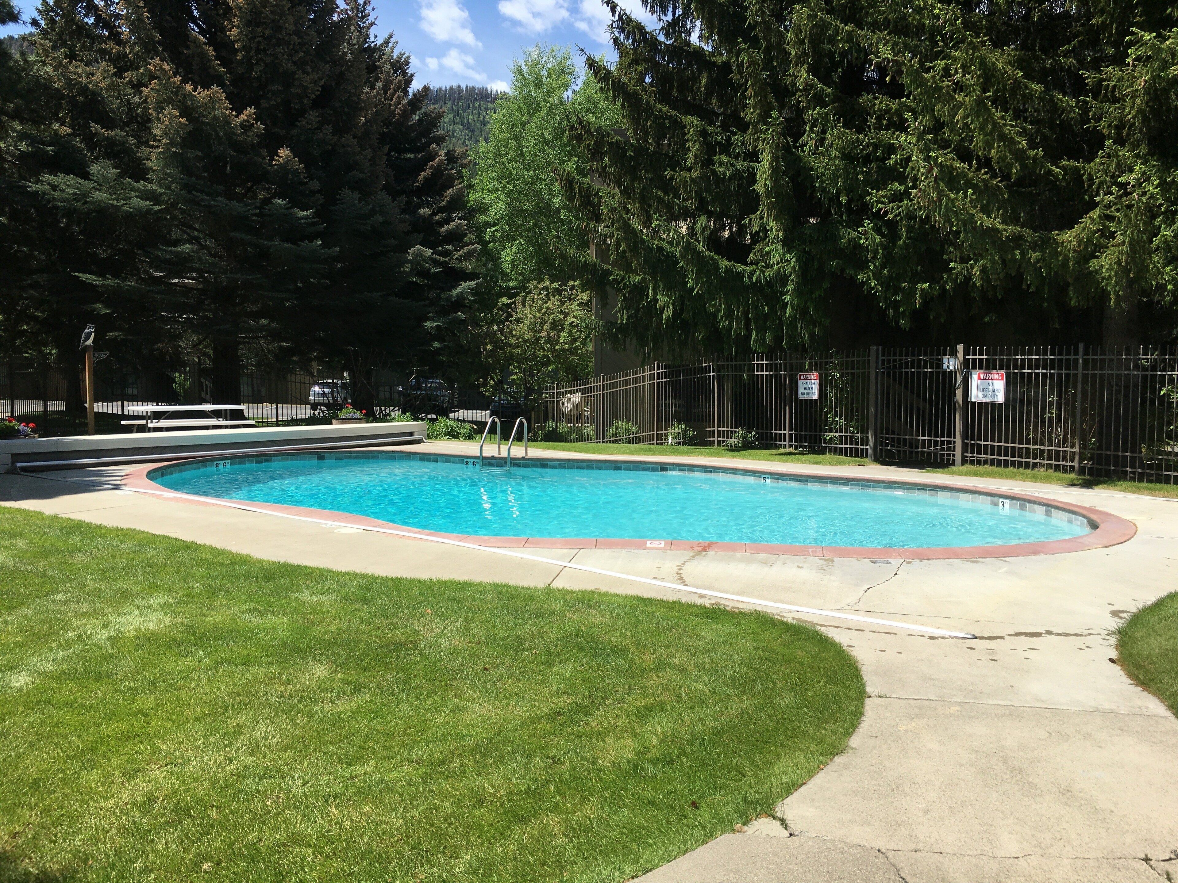 A rectangular swimming pool with a surrounding deck, a lifeguard sign, and a fenced area with trees.