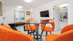 Uma sala moderna com uma mesa de jantar de vidro, cadeiras laranja, um sofá, uma TV fixada na parede e uma porta deslizante que dá para um banheiro.