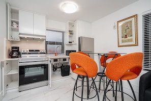 A compact kitchen with white cabinets, a stainless steel refrigerator, a built-in oven, and a coffee maker. There are two orange bar stools and a small table. A framed picture of a rose is on the wall. - Vancouver Brand New 2Beds 2 Full Baths 1 Sofa Bed Laneway House,Steps 2 SkyTrain (Vancouver)