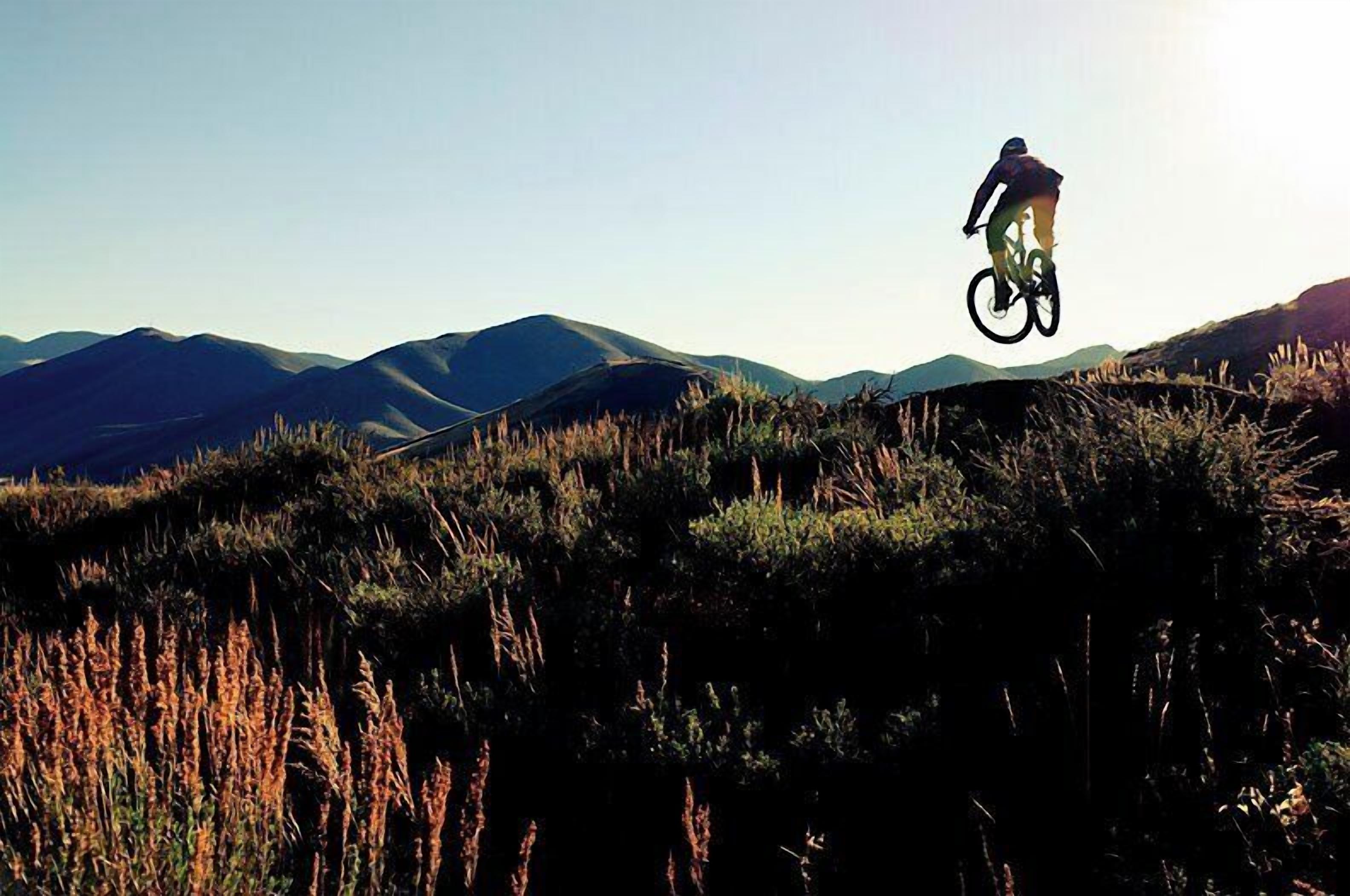 A person on a mountain bike performing a jump over a grassy field with mountains in the background.