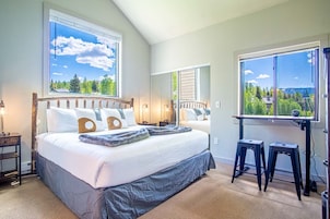 Quarto com uma cama grande, duas janelas com vista para vegetação, uma escrivaninha com dois banquinhos e uma mesa de cabeceira com abajur.