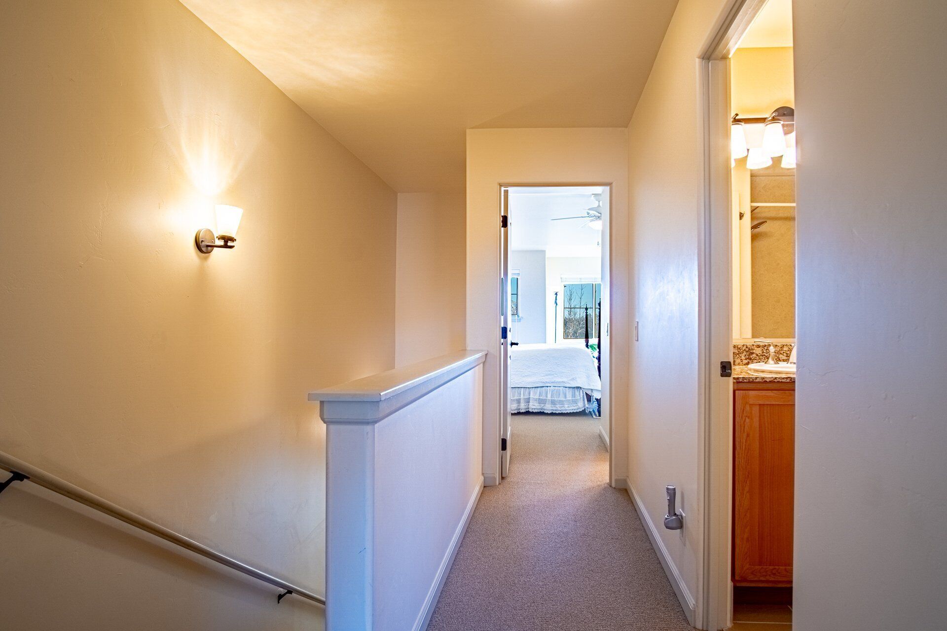 A narrow hallway with a carpeted floor, a white railing, and a light fixture on the left wall.