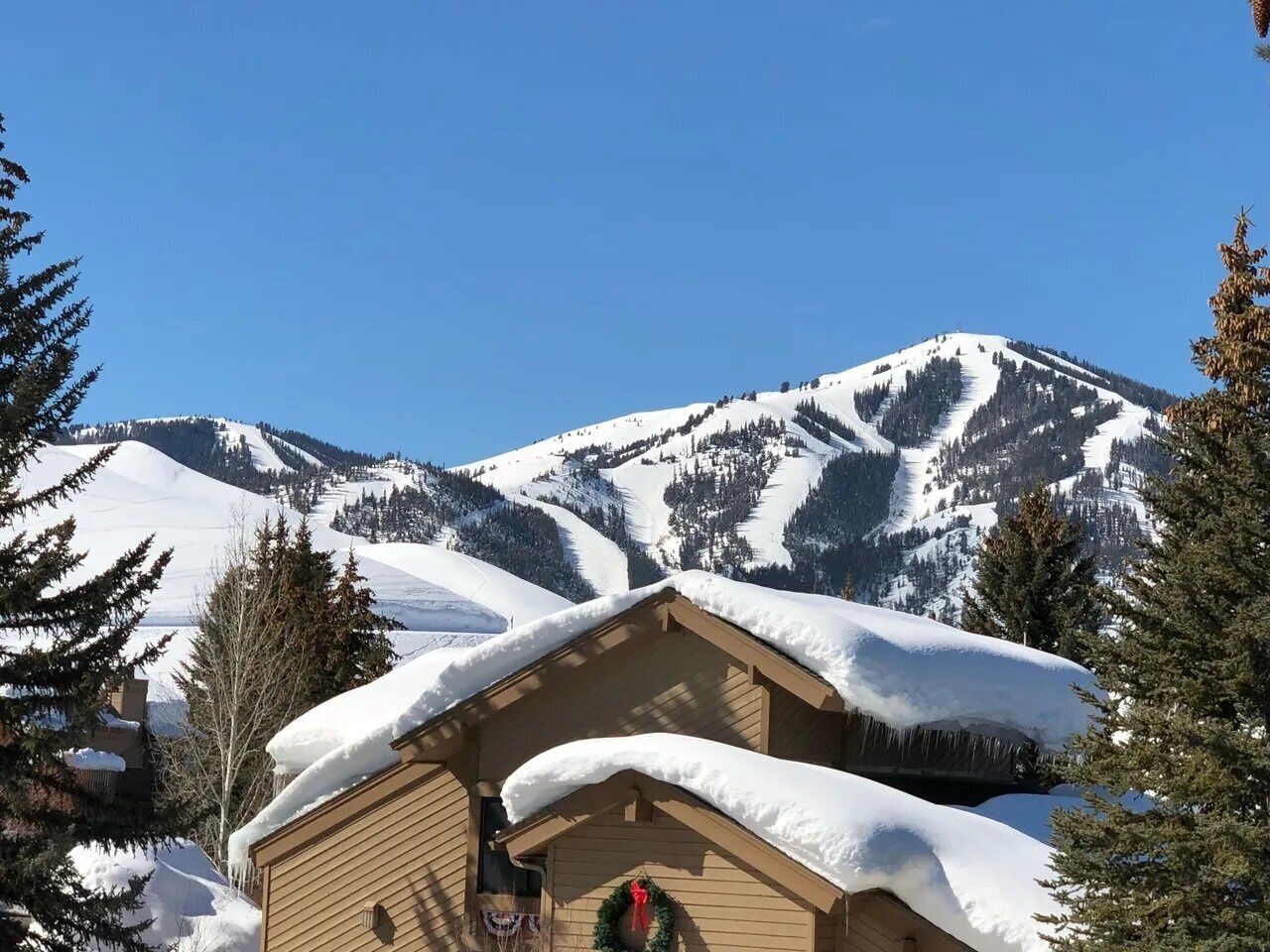 Una baita di legno con tetto imbiancato dalla neve e una ghirlanda sulla porta, circondata da alberi e montagne innevate.