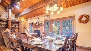 Zona pranzo con un tavolo in legno apparecchiato per quattro persone, circondato da sedie. Si nota un camino con l'orologio sopra, e una ghirlanda appesa al muro.