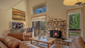 A living room with a stone fireplace, leather couches, a television, and a large window. - Bonne Vie Condo - Heart of Elkhorn + Resort Amenities (Sun Valley)