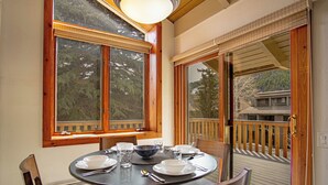 Un coin repas avec une table dressée pour quatre personnes, une fenêtre donnant sur la forêt et une terrasse en bois à l’extérieur.