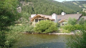 Il s’agit d’un chalet doté d’une façade en pierre, de grandes fenêtres et d’une cheminée. Il est entouré d’une forêt dense et bordé par une rivière.