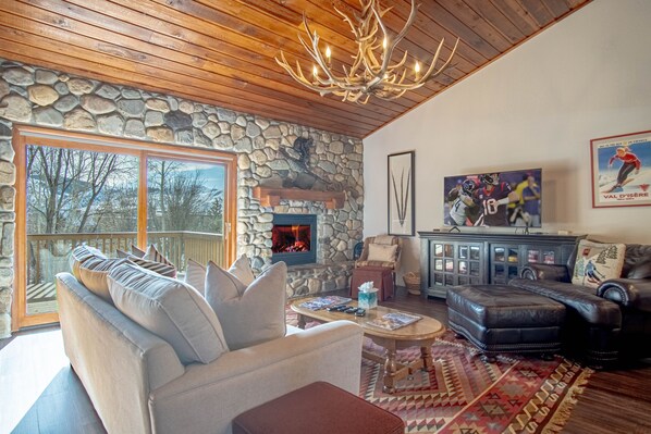 A cozy living room with a stone wall, a fireplace, a large window, and a chandelier. - Ridge Condo - Modern Luxury Condo W/resort Amenities (Sun Valley)