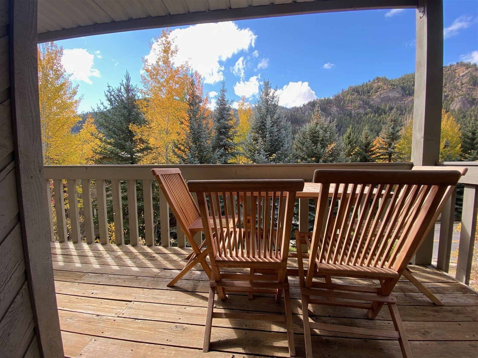 A wooden deck with two wooden chairs and a table, overlooking a forested hillside with autumn foliage.