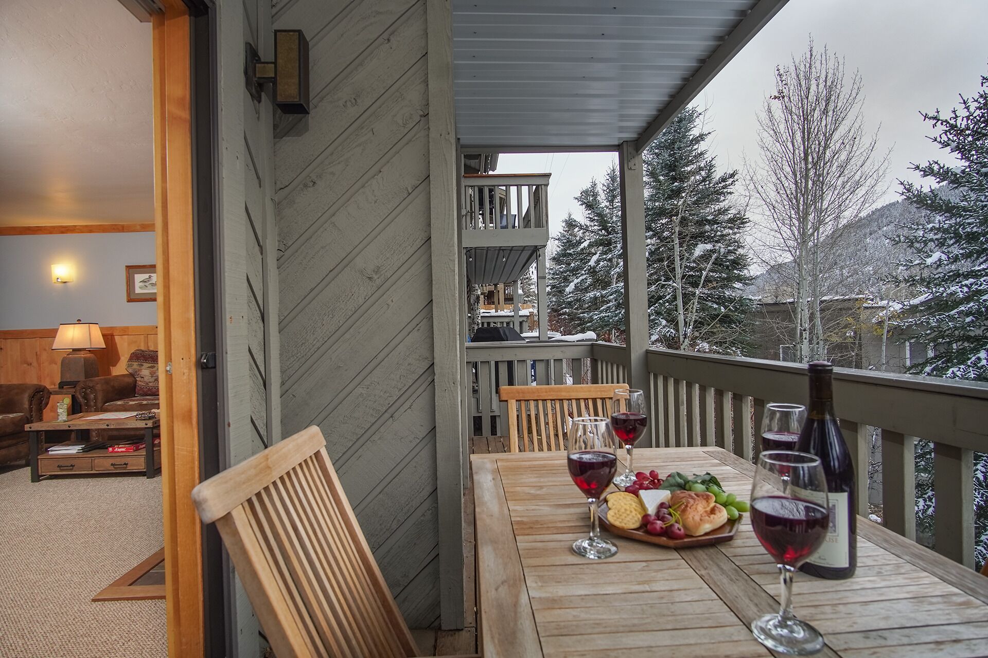 Eine Holzterrasse mit Essplatz, Wein gläsern und einer Flasche, mit Blick auf eine verschneite Landschaft mit Bäumen.