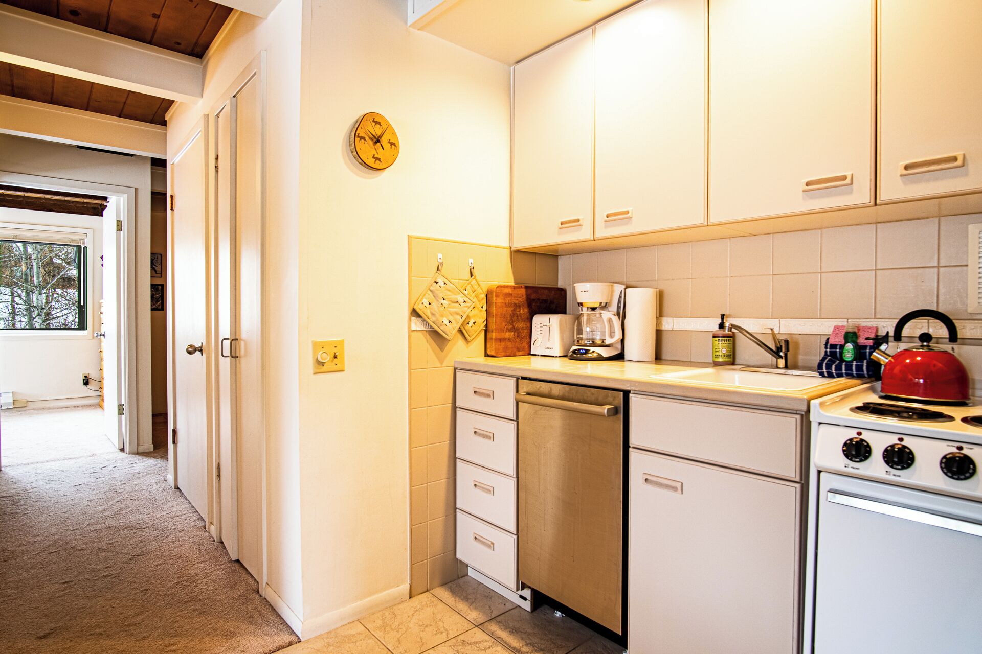Una cocina con gabinetes blancos, cafetera, hervidor y un reloj en la pared.