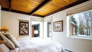 Un dormitorio con techo de madera, un ventanal con vistas a árboles, una cama con ropa blanca y dos obras de arte colgadas en la pared.