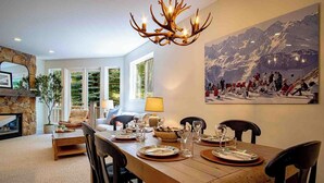 A dining area with a wooden table set for four, surrounded by chairs. A chandelier with antler-like arms hangs above. - Snow Star Serenade - Hot Tub + Gym + Walk to Downtown & ski Lifts! (Ketchum)
