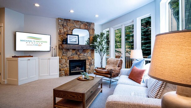 A modern living room with a stone fireplace, a television mounted on the wall, and a large window offering a view of the outdoors. - Snow Star Serenade - Hot Tub + Gym + Walk to Downtown & ski Lifts! (Ketchum)