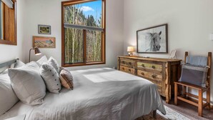 Quarto com uma janela grande, cómoda de madeira, cadeira e cama com almofadas.