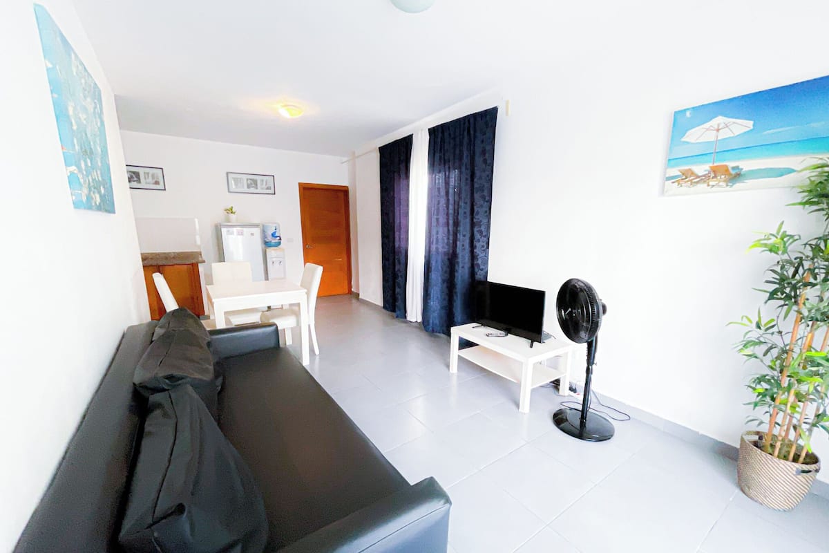 Una sala moderna con un sofá de cuero negro, una mesa de centro blanca, un televisor de pantalla plana y un ventilador de pie. Hay un pequeño comedor con una mesa y sillas blancas, y una zona de cocina con un refrigerador y dispensador de agua. Se ve una ventana grande con cortinas oscuras, y una planta en maceta está colocada cerca de la ventana.
