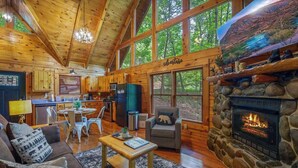 A log cabin interior with a stone fireplace, wooden furniture, a kitchen area, and large windows. - Secluded Cabin for 4 w/ Private Hot Tub & Jacuzzi! (Sevierville)