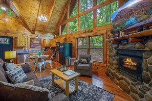 A log cabin interior with a stone fireplace, wooden furniture, a kitchen area, and large windows. - Secluded Cabin for 4 w/ Private Hot Tub & Jacuzzi! (Sevierville)