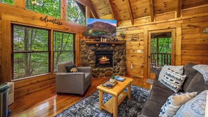 A log cabin interior with a stone fireplace, a gray sofa, a wooden coffee table, and large windows. - Secluded Cabin for 4 w/ Private Hot Tub & Jacuzzi! (Sevierville)