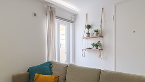 Una sala de estar en tonos neutros, con un sofá color beige, un cojín amarillo, una manta verde azulado y una estantería de madera para plantas.