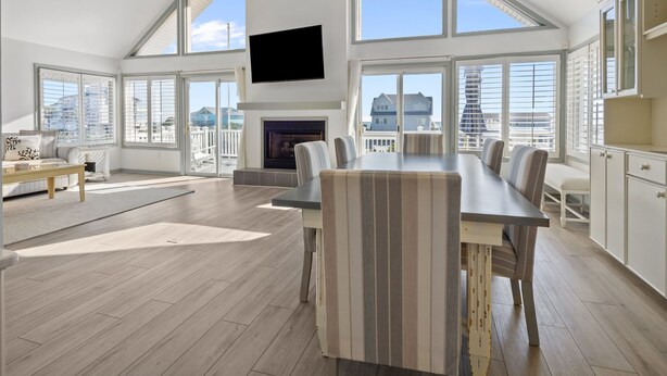 A spacious living room with a high ceiling, large windows, a fireplace, and a flat-screen TV mounted on the wall. - Steps to beach with views galore! (Emerald Isle)