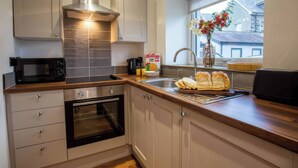A modern kitchen with white cabinets, a stainless steel range hood, a microwave, and a window with a view of buildings. - Aspen Apartment (Llanrwst)