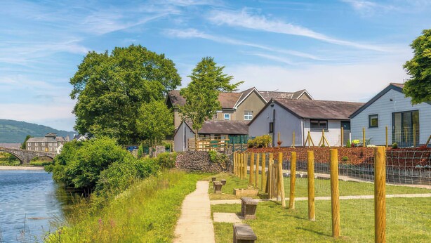 A riverside pathway with stone benches, a wooden fence, and houses with sloped roofs. - Aspen Apartment (Llanrwst)