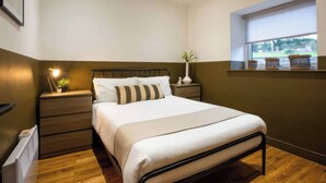 A bedroom with a bed, bedside tables, a nightstand, a window with a view, and a wall-mounted shelf with baskets. - Aspen Apartment (Llanrwst)