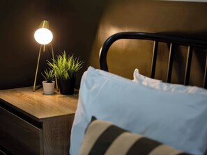 A neatly made bed with a striped pillow, a bedside table with a lamp and two potted plants, and a dark headboard. - Aspen Apartment (Llanrwst)