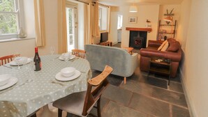 A dining area with a round table, chairs, and a bottle of wine. There is a sofa and a fireplace in the background. - Rock Cottage (Milnthorpe)