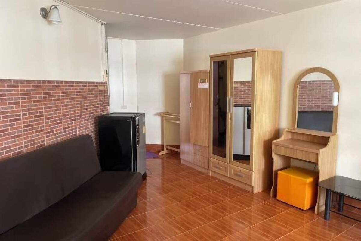 Una habitación con un sofá de color oscuro, un armario de madera, un taburete pequeño naranja y suelo de baldosas.