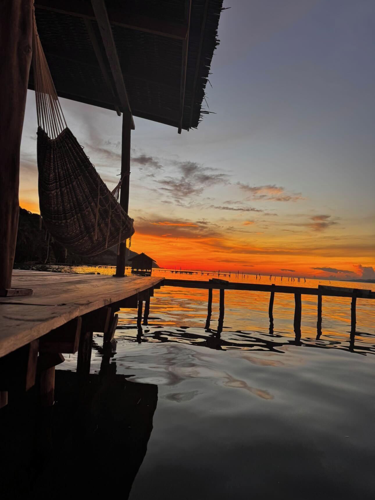 A wooden pier extending into calm waters at sunset, with a thatched-roof structure on the left.