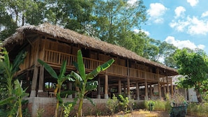 A wooden structure with a thatched roof, surrounded by greenery and trees. - Suoi Rao Hao Huc Village (Ho Chi Minh City)