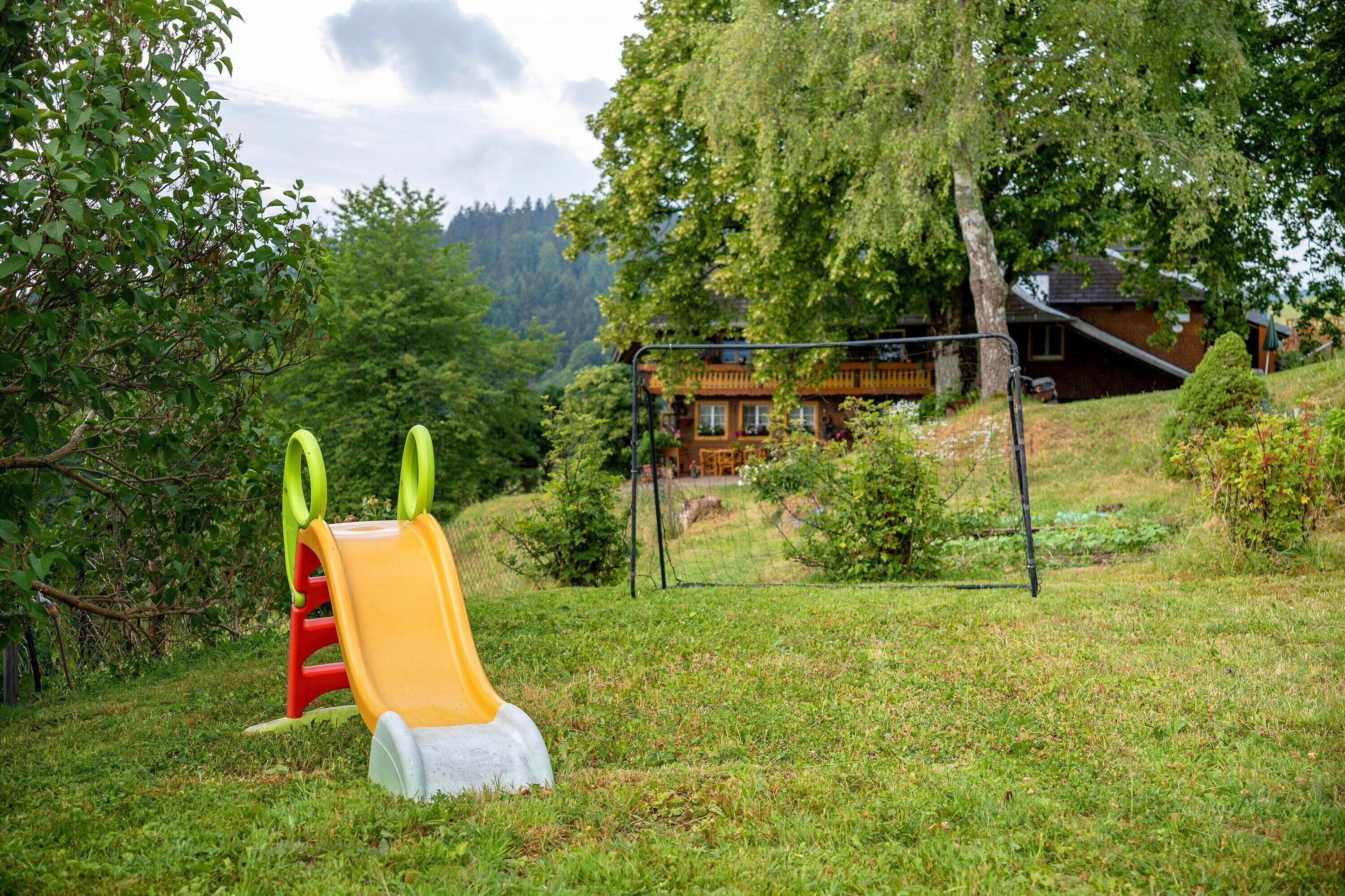 Ein Spielplatz mit Rutsche und Schaukeln, umgeben von Bäumen und einem Haus im Hintergrund.