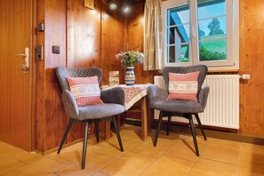 Une pièce aux murs lambrissés, avec deux fauteuils gris à revêtement en tissu, une petite table sur laquelle est posé un vase de fleurs et une fenêtre donnant sur un paysage verdoyant.