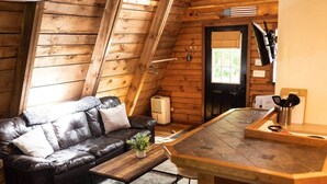 A cozy cabin interior with a leather sofa, a wooden dining table, and a small kitchen area. - Canoe Believe It Cabin | Indoor Jacuzzi Tub (Powell County)