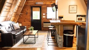 A cozy log cabin interior with a black leather sofa, a wooden coffee table, a kitchen area with a bar counter, and a stone sink. - Canoe Believe It Cabin | Indoor Jacuzzi Tub (Powell County)