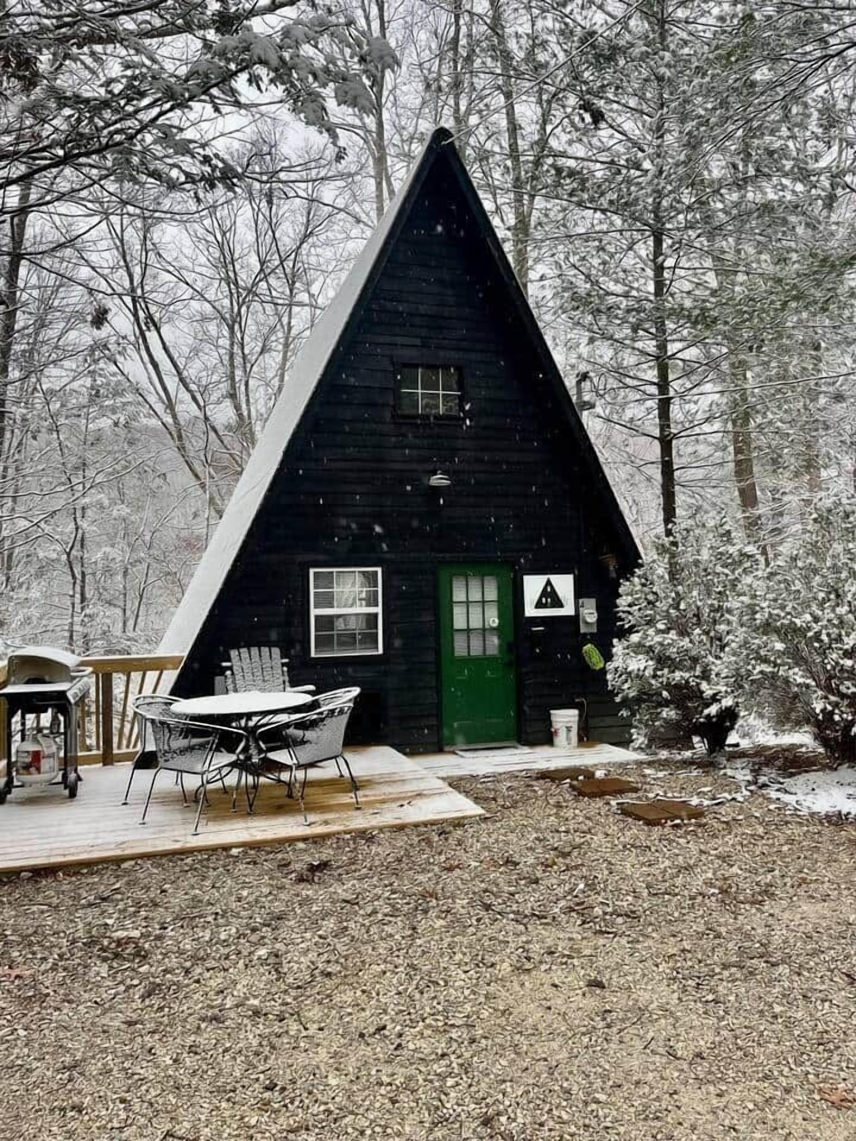 A triangular cabin with a green door and windows, surrounded by trees in a snowy landscape.